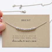 Hand holding the Extra Bold sterling silver Morse code necklace on its card, the name Brent spelled in Morse code beads