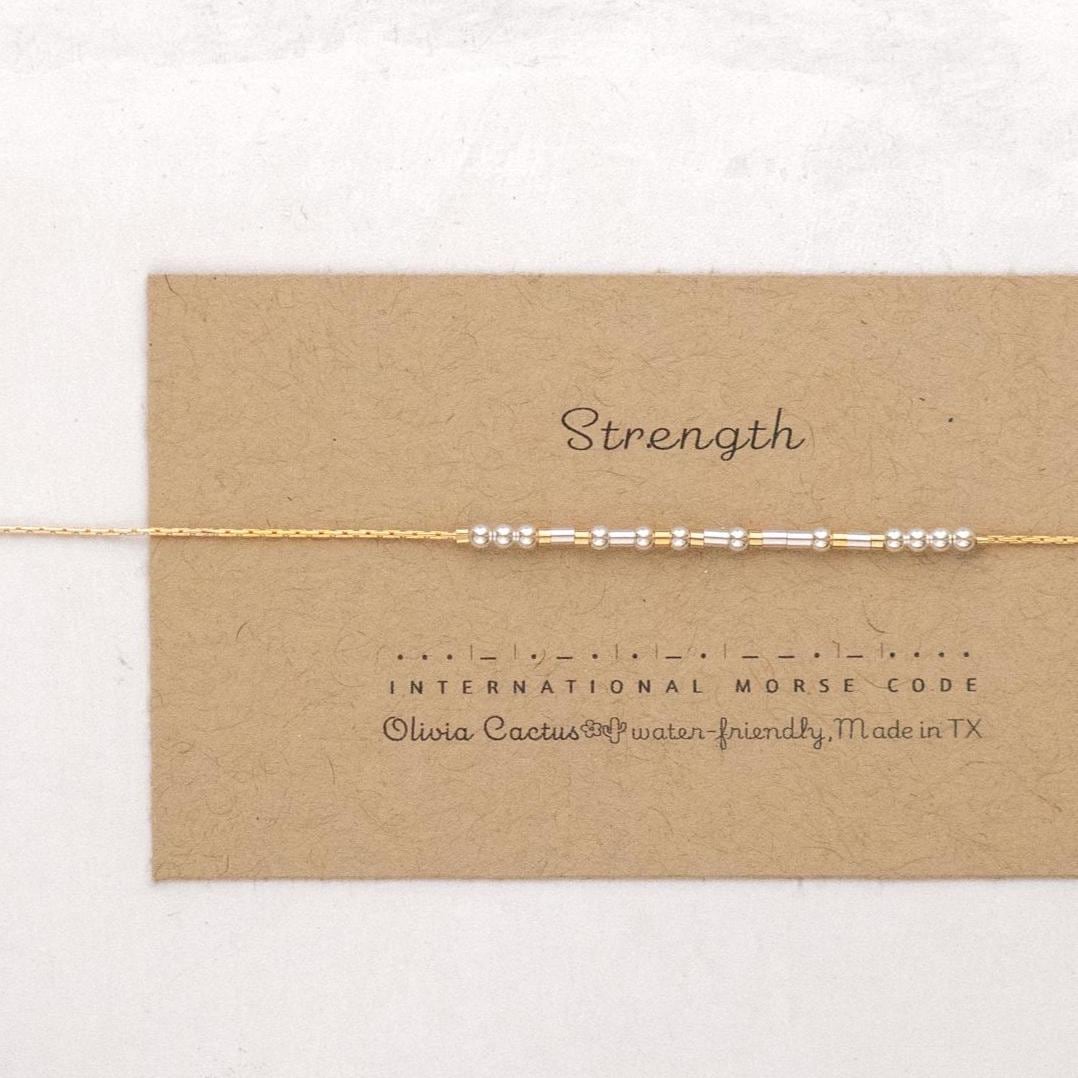 May include: A gold chain bracelet with silver beads spelling out the word &amp;quot;Strength&amp;quot; in International Morse Code. The bracelet is on a brown card with the text &amp;quot;Olivia Cactus® Water-Friendly, Made in TX&amp;quot;.