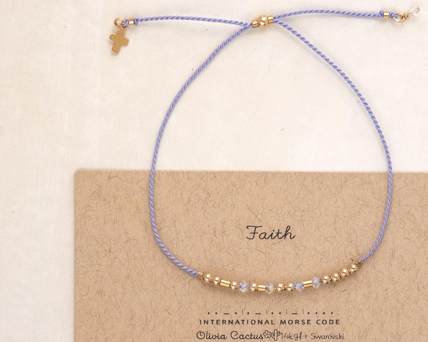 May include: A delicate bracelet with a light purple cord and gold-colored beads. A small gold cross charm hangs from the cord. The bracelet is displayed on a card that reads "Faith" in cursive.