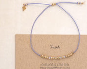 May include: A delicate bracelet with a light purple cord and gold-colored beads. A small gold cross charm hangs from the cord. The bracelet is displayed on a card that reads "Faith" in cursive.