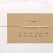 May include: A gold chain bracelet with silver beads spelling out the word "Strength" in International Morse Code. The bracelet is on a brown card with the text "Olivia Cactus® Water-Friendly, Made in TX".